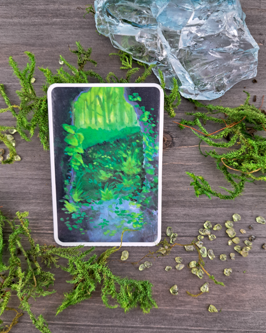 An image of a sticker surrounded by crystals and moss. The sticker features a vertical landscape with foliage flowing into a cave exit (portal) as you look outside from it, which was painted with acrylic inks.