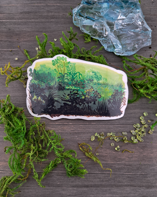 An image of a sticker surrounded by crystals and moss. The sticker features a horizontal landscape with a variety of foliage and green gradients, which was painted with acrylic inks.