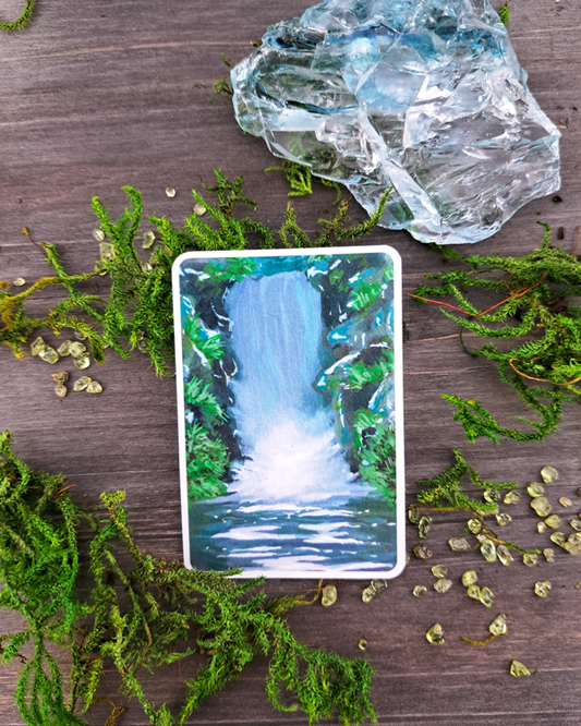 An image of a sticker surrounded by crystals and moss. The sticker features a vertical landscape with foliage and a waterfall, which was painted with acrylic inks.