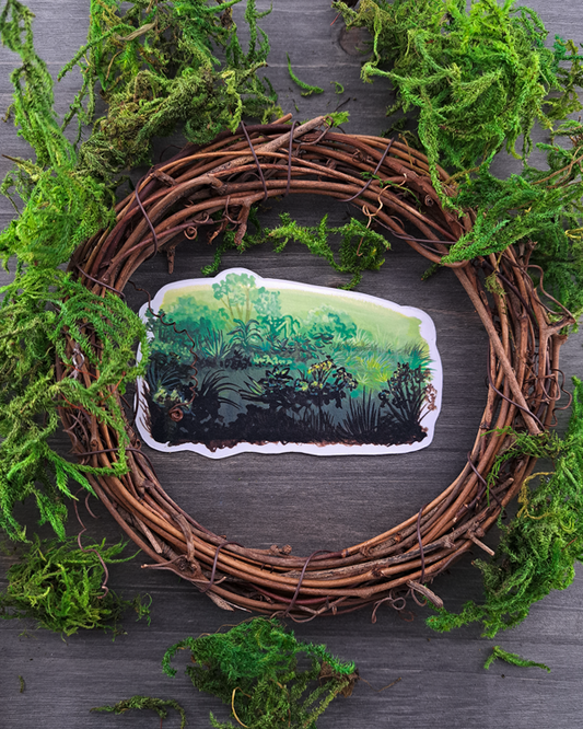 An image of a sticker surrounded by a wooden wreath and moss. The sticker features a horizontal landscape with a variety of foliage and green gradients, which was painted with acrylic inks.