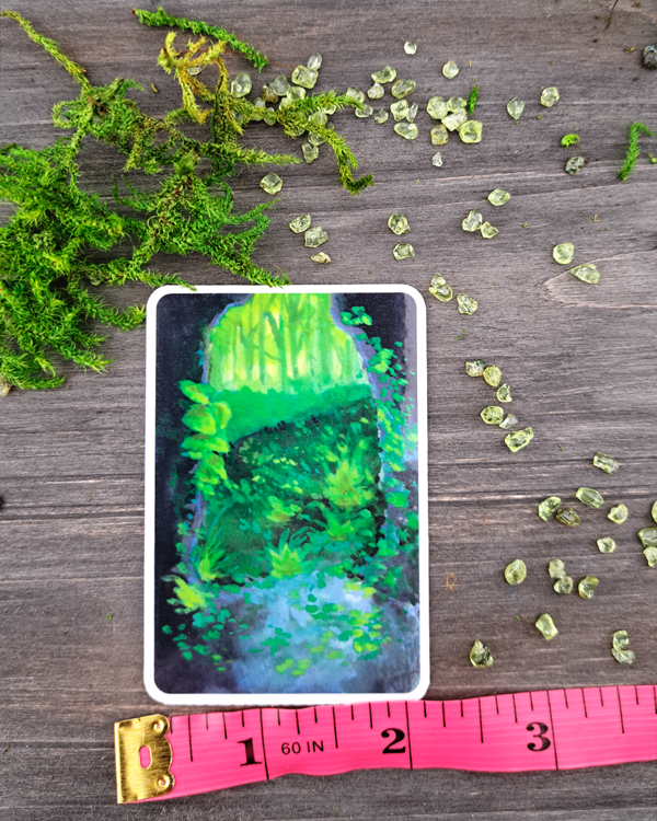An image of a sticker surrounded by crystals and moss, with a pink ruler underneath to show size. The sticker features a vertical landscape with foliage flowing into a cave exit (portal) as you look outside from it, which was painted with acrylic inks.