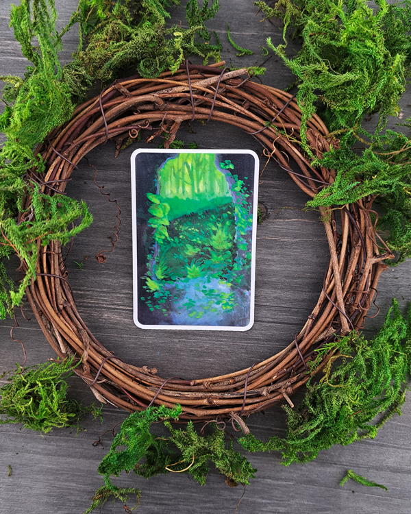 An image of a sticker surrounded by a wooden wreath and moss. The sticker features a vertical landscape with foliage flowing into a cave exit (portal) as you look outside from it, which was painted with acrylic inks.