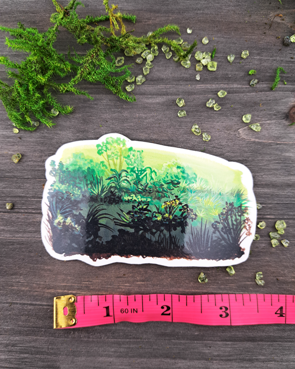 An image of a sticker surrounded by crystals and moss and a pink ruler underneath to show size. The sticker features a horizontal landscape with a variety of foliage and green gradients, which was painted with acrylic inks.