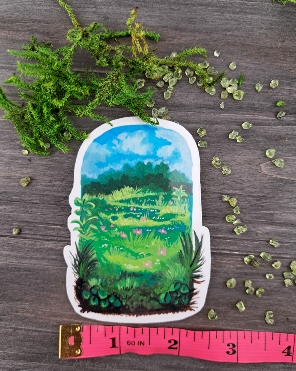 An image of a sticker surrounded by crystals and moss and a pink ruler underneath. The sticker features a vertical landscape with foliage in a meadow that contains trees, grass pink flowers and a blue sky with clouds, which was painted with acrylic inks.