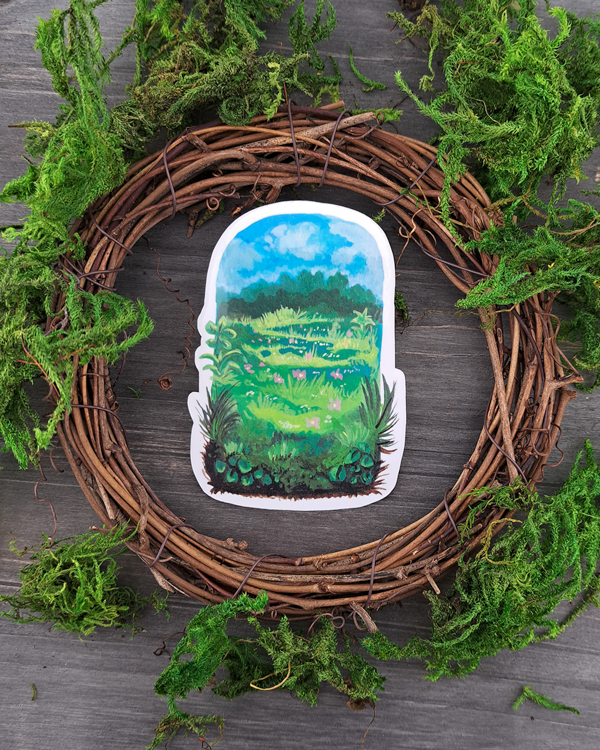 An image of a sticker surrounded by a wooden wreath and moss. The sticker features a vertical landscape with foliage in a meadow that contains trees, grass pink flowers and a blue sky with clouds, which was painted with acrylic inks.