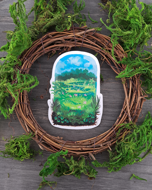 An image of a sticker surrounded by a wooden wreath and moss. The sticker features a vertical landscape with foliage in a meadow that contains trees, grass pink flowers and a blue sky with clouds, which was painted with acrylic inks.