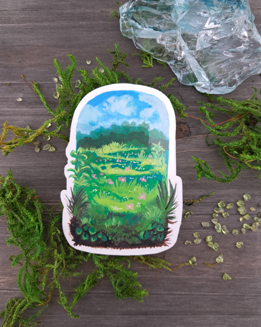 An image of a sticker surrounded by crystals and moss. The sticker features a vertical landscape with foliage in a meadow that contains trees, grass pink flowers and a blue sky with clouds, which was painted with acrylic inks.