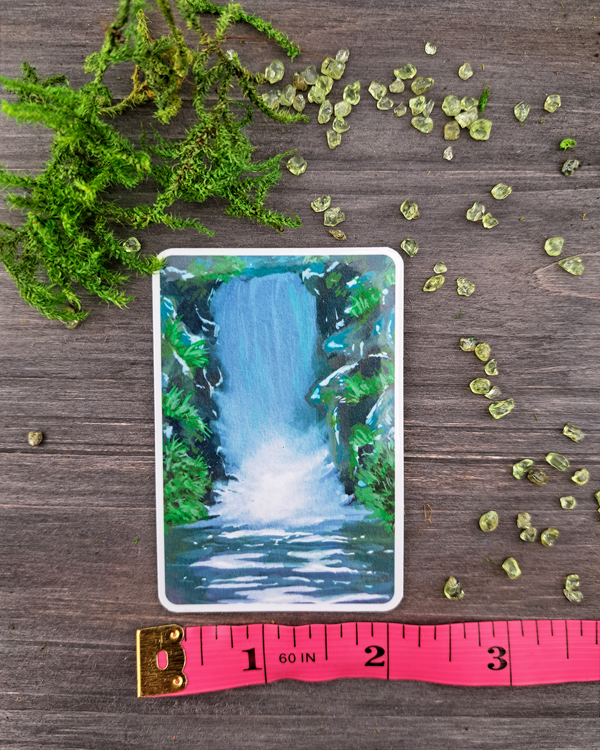 An image of a sticker surrounded by green crystals and moss. The sticker features a vertical landscape with foliage and a waterfall, which was painted with acrylic inks.