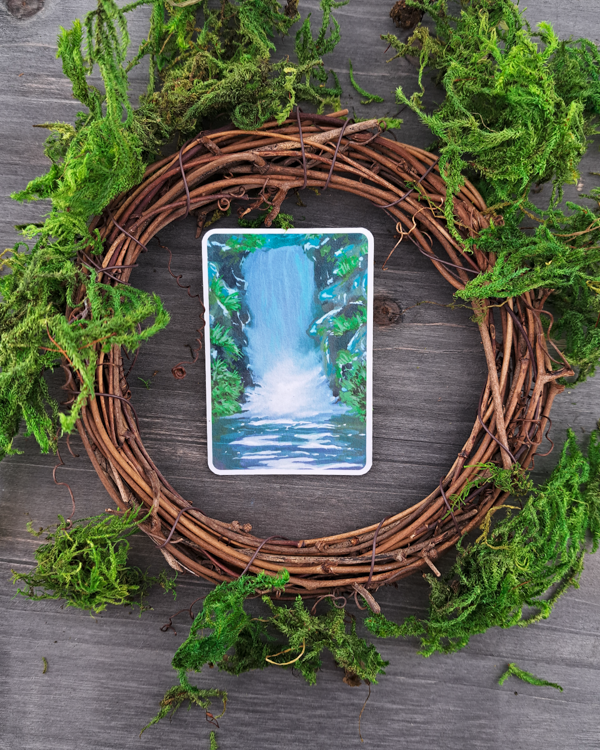 An image of a sticker surrounded by a wooden wreath and moss. The sticker features a vertical landscape with foliage and a waterfall, which was painted with acrylic inks.
