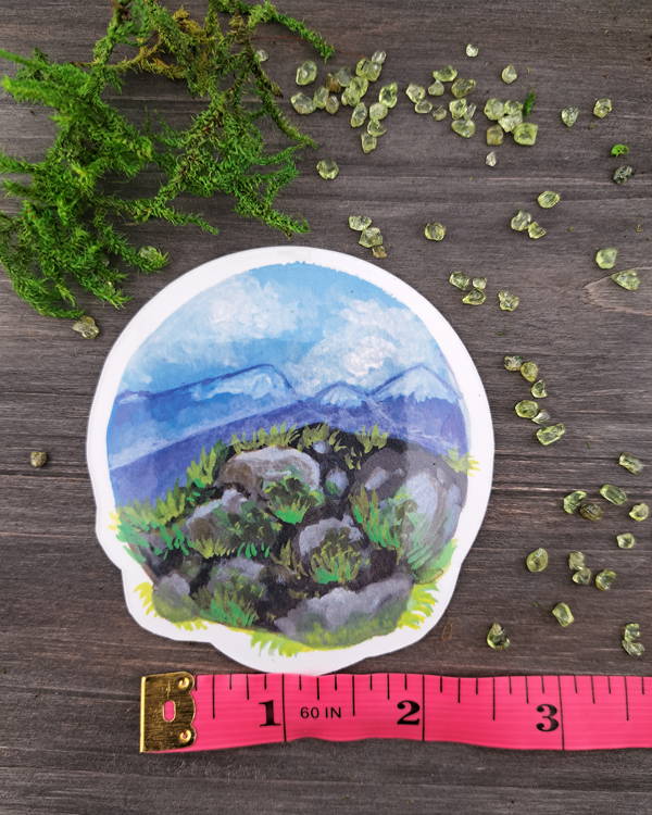 An image of a sticker surrounded by crystals and moss. The sticker features a round landscape with rocks and foliage overlooking a mountain valley, which was painted with acrylic inks.