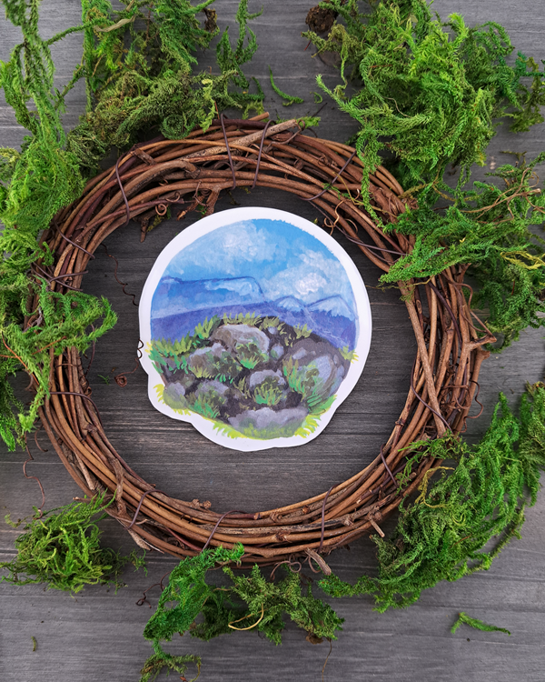 An image of a sticker surrounded by a wooden wreath and moss. The sticker features a round landscape with rocks and foliage overlooking a mountain valley, which was painted with acrylic inks.