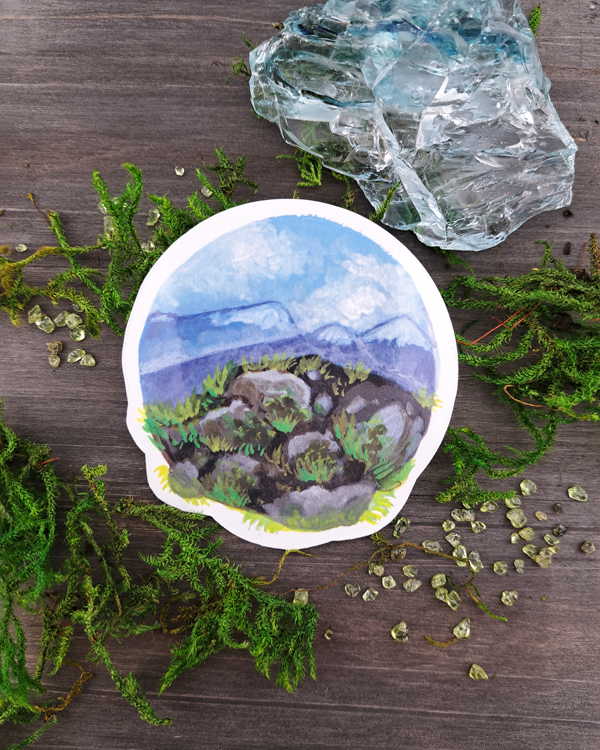 An image of a sticker surrounded by crystals and moss. The sticker features a round landscape with rocks and foliage overlooking a mountain valley, which was painted with acrylic inks.