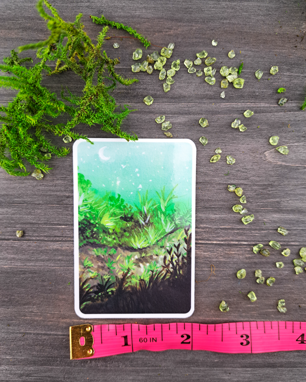 An image of a sticker surrounded by crystals and moss, and a pink ruler underneath to show size. The sticker features a vertical landscape with foliage along a pathway with a white moon shown in the pale blue sky, which was painted with acrylic inks.