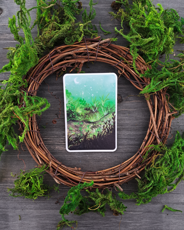 An image of a sticker surrounded by a wooden wreath and moss. The sticker features a vertical landscape with foliage along a pathway with a white moon shown in the pale blue sky, which was painted with acrylic inks.