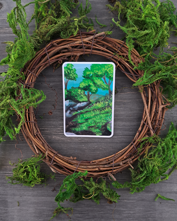 An image of a sticker surrounded by a wooden wreath and moss. The sticker features a landscape with rocks, trees and grass, which was painted with acrylic inks.