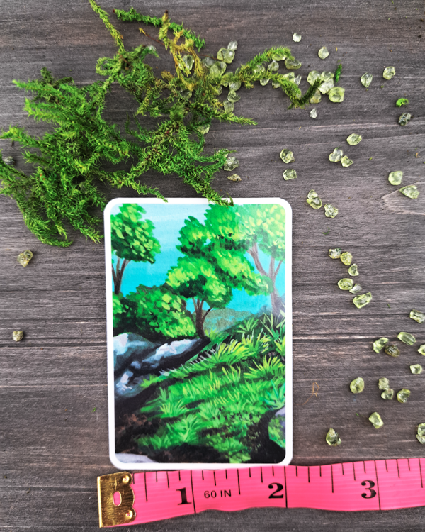 An image of a sticker surrounded by green crystals and moss with a pink ruler underneath to show the size. The sticker features a landscape with rocks, trees and grass, which was painted with acrylic inks.