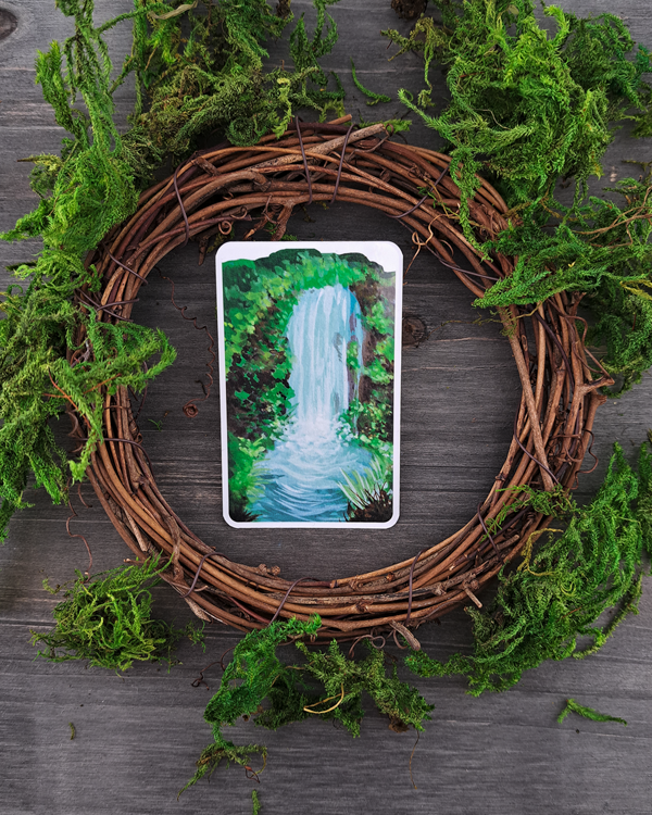 An image of a sticker surrounded by a wooden wreath and moss. The sticker features a vertical landscape with foliage and a waterfall, which was painted with acrylic inks.