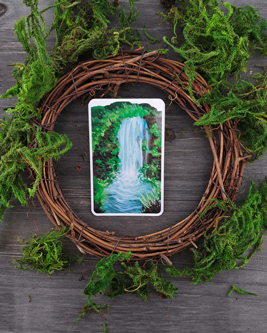 An image of a sticker surrounded by a wooden wreath and moss. The sticker features a vertical landscape with foliage and a waterfall, which was painted with acrylic inks.