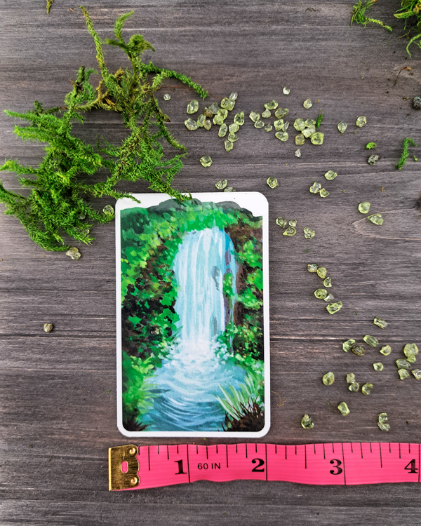 An image of a sticker surrounded by crystals and moss and a pink ruler below to show size. The sticker features a vertical landscape with foliage and a waterfall, which was painted with acrylic inks.