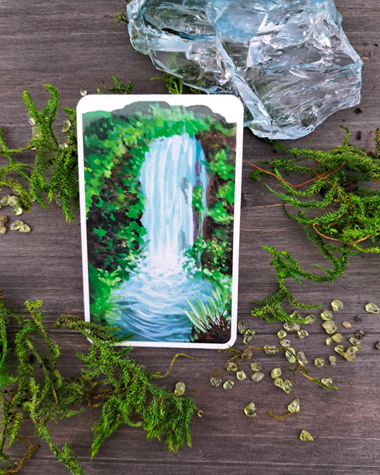 An image of a sticker surrounded by crystals and moss. The sticker features a vertical landscape with foliage and a waterfall, which was painted with acrylic inks.